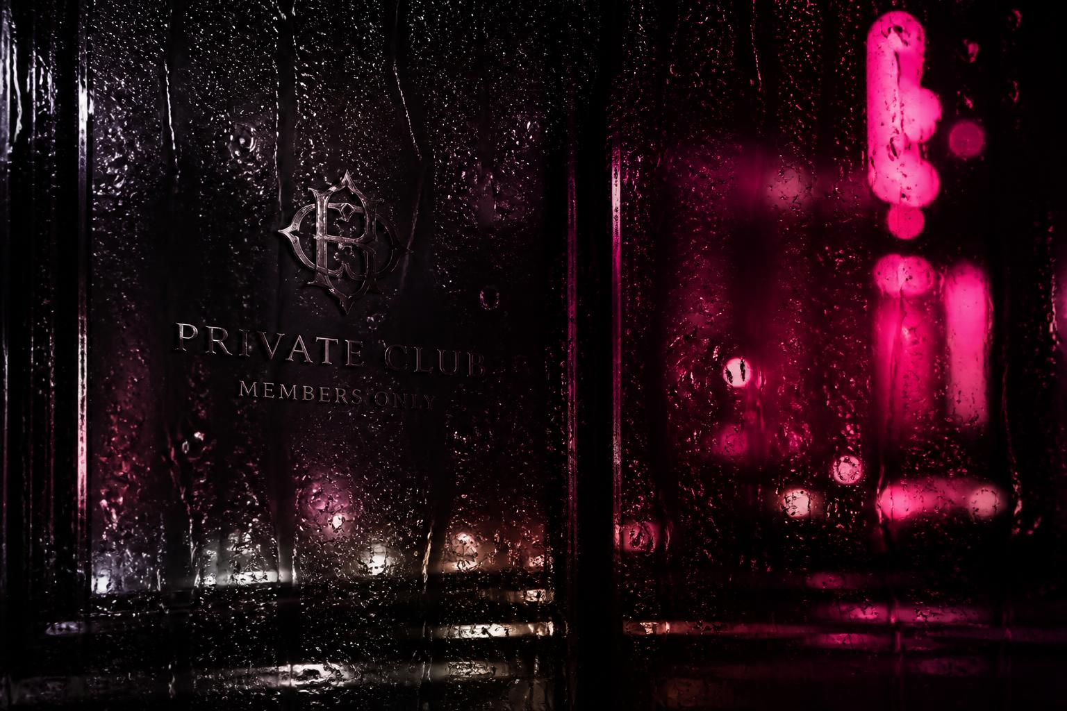 Rain streaming down dark glass outside a private club at night